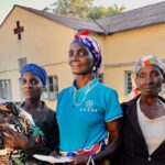 Three bold Angolan women, one holding an infant, standing in front of the Kalukembe waiting home